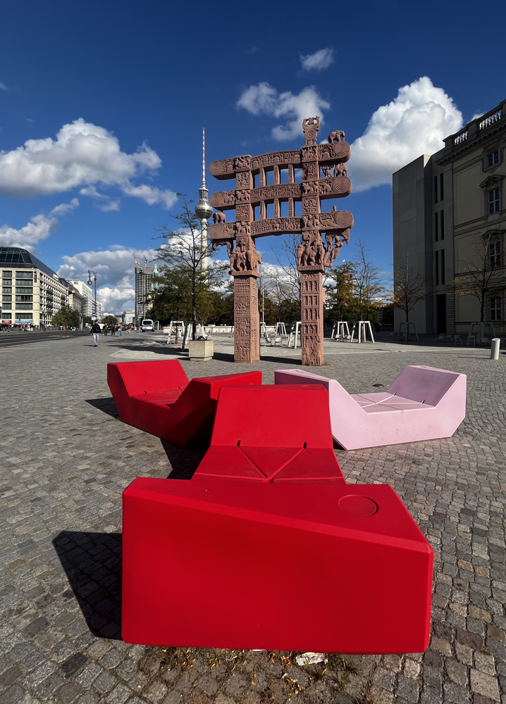 HUMBOLT FORUM -  repliKA istočne kapije (Torana) Velike stupe u Sanchiju, drevnog budističkog spomenika u Indiji. Ova se replika nalazi ispred Humboldt Foruma u Berlinu. Materijal: Iako izgleda kao da je drvena, izvorna stupa u Sanchiju i njezine kapije izrađene su od pje&scaron;čenjaka, a i ova je replika napravljena od pje&scaron;čenjaka.   Nalazi se na trgu ispred Humboldt Foruma, a u pozadini fotografije vidi se berlinski TV toranj.   Izvorna kapija datira iz 1. stoljeća prije Krista. Gipsani odljevci originala doneseni su u Berlin krajem 19. stoljeća, a ova nova replika dovr&scaron;ena je 2020-ih.   Složeni reljefi prikazuju prizore iz Buddhinog života i Jataka priča. Original se smatra jednom od najstarijih i najvažnijih struktura budističke arhitekture.  &Scaron;areni objekti u prednjem planu moderne su instalacije za sjedenje, vjerojatno izrađene od  plastike.